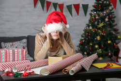 Women stressed and covering her eyes with a Santa hat while wrapping presents.